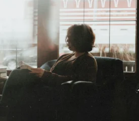 A person sits in a cozy chair near a large window, reading a book as soft daylight filters into the room