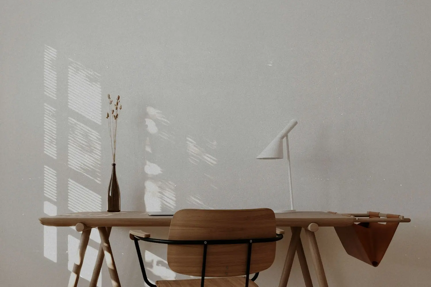 andinavian-style workspace featuring a wooden desk, simple chair, white lamp, and dried botanicals with sunlight filtering through a nearby window