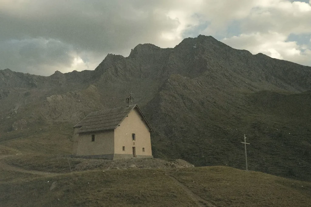 A small church sits on a grassy hillside beneath towering, rugged mountains under a cloudy sky