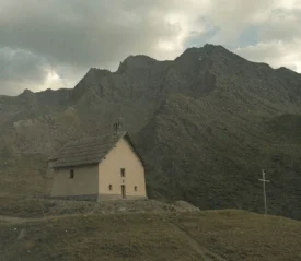 A small church sits on a grassy hillside beneath towering, rugged mountains under a cloudy sky