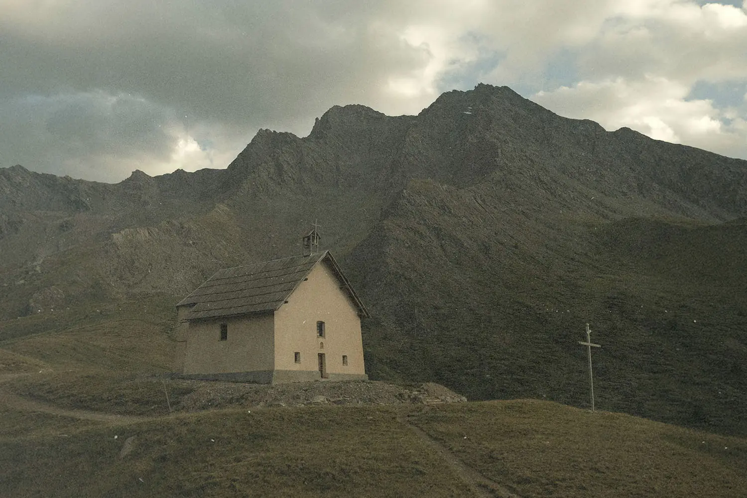 A small church sits on a grassy hillside beneath towering, rugged mountains under a cloudy sky