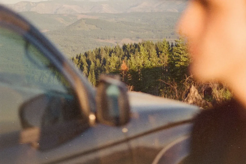 Parked car is shown in the foreground while a scenic landscape of rolling hills and evergreen forests stretches into the distance