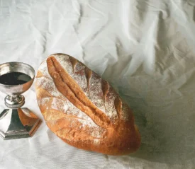 A loaf of rustic bread rests beside a silver chalice filled with dark liquid on a light, textured cloth