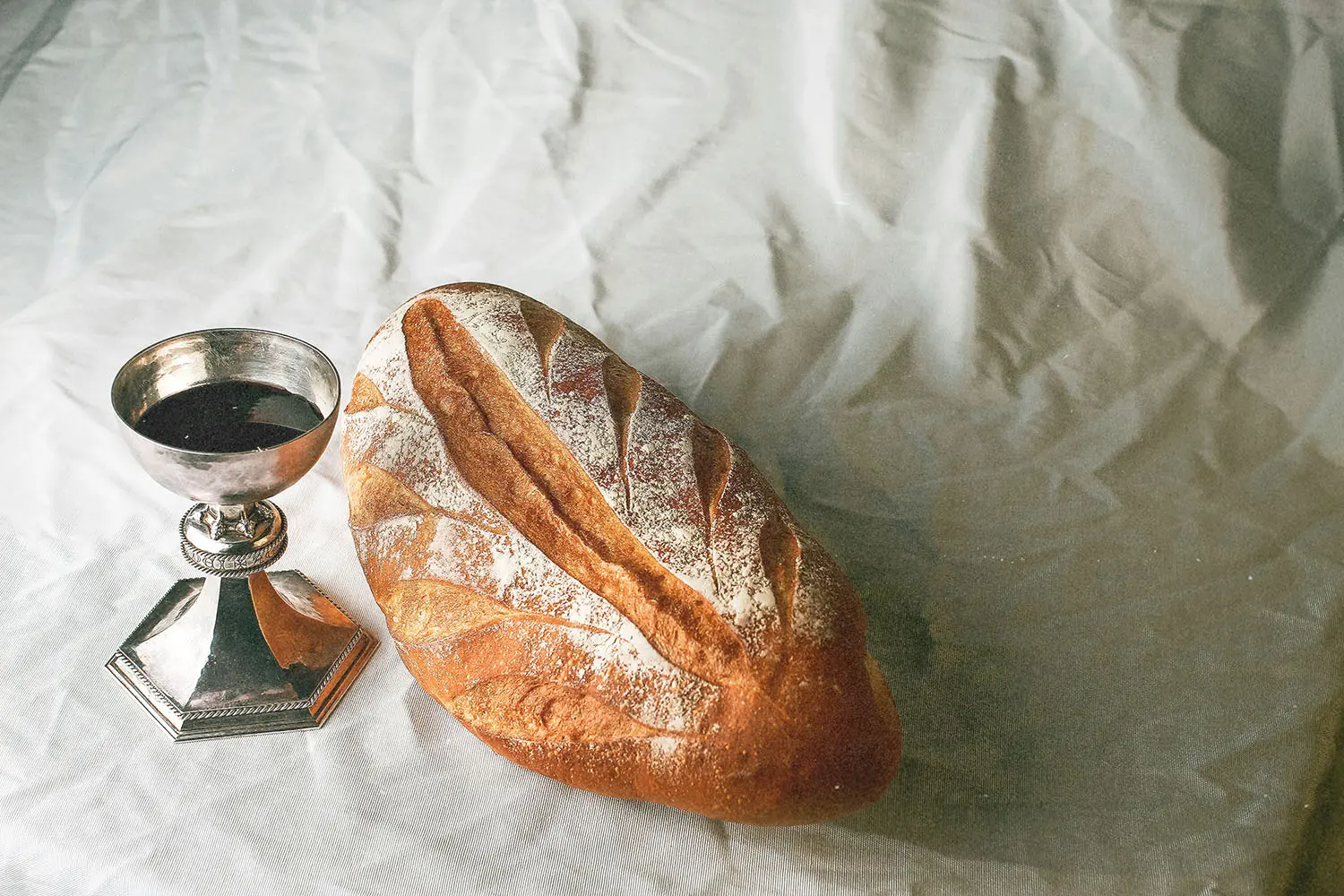A loaf of rustic bread rests beside a silver chalice filled with dark liquid on a light, textured cloth