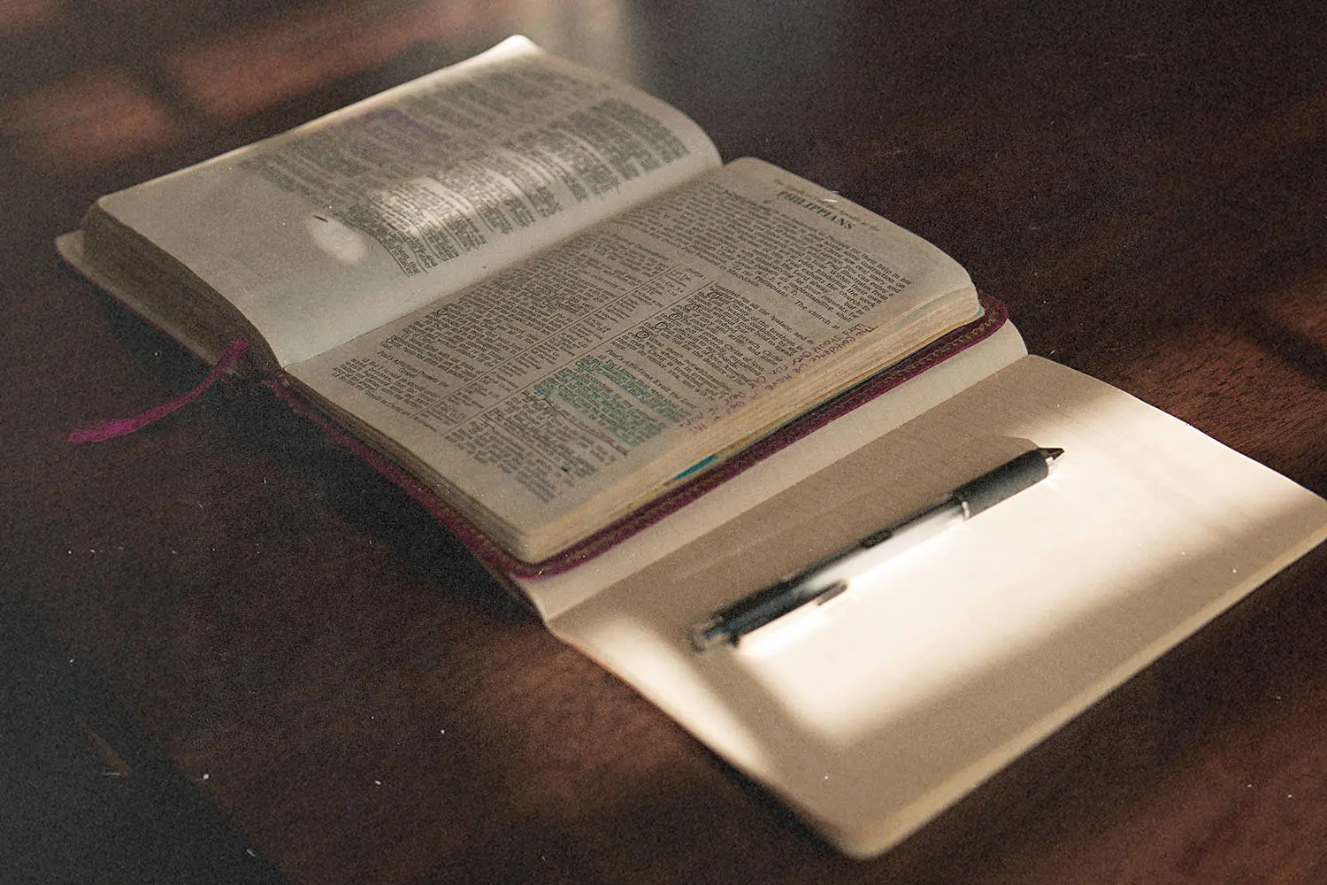 An open Bible rests on a table beside a blank notebook and pen, lit by soft sunlight from a nearby window