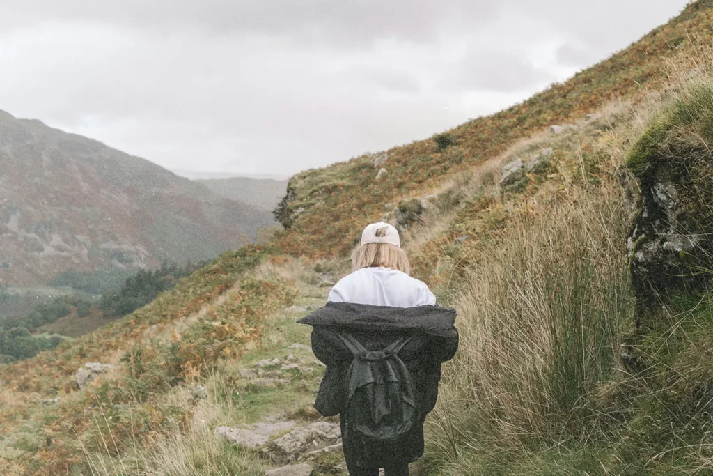 A person walks alone along a narrow mountain trail, wearing a light jacket and backpack as misty hills rise on both sides