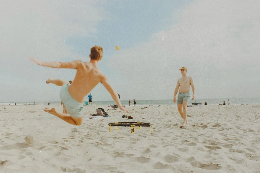 Two people on a beach playing spike ball, as one jumps into the air to hit the ball over the sand on a sunny shoreline