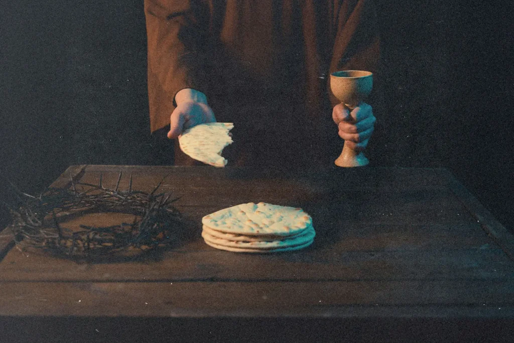 Hands holding unleavened bread and a communion cup above a wooden table with matzo and a crown of thorns