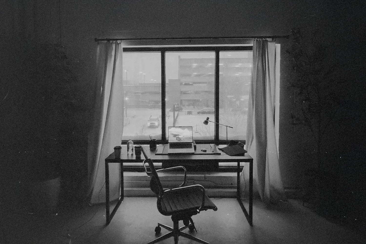 Simple office space with a laptop and lamp on a desk framed by open curtains and an urban view