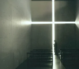 A person stands inside a dimly lit chapel, silhouetted against a large glowing cross-shaped opening on the far wall