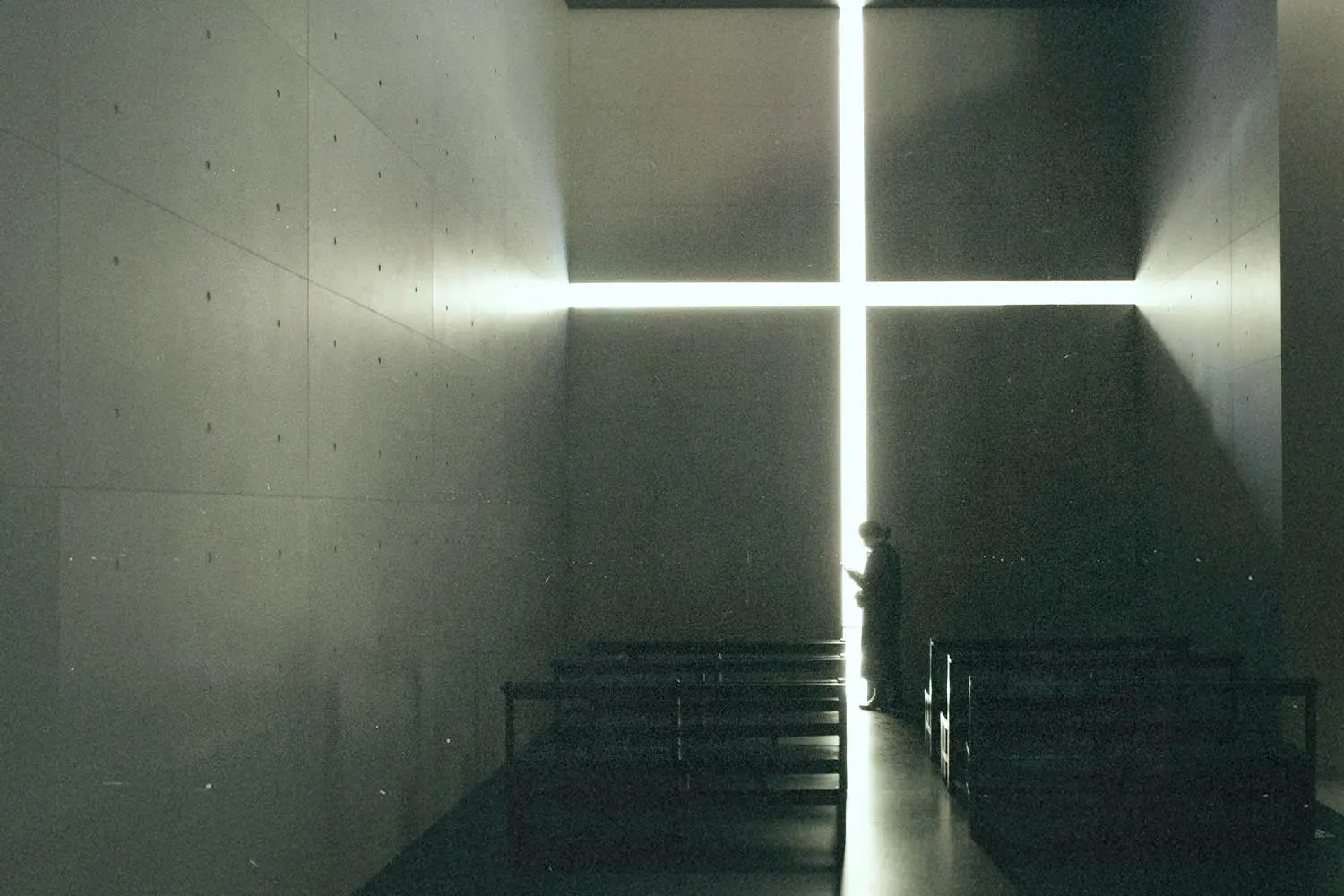 A person stands inside a dimly lit chapel, silhouetted against a large glowing cross-shaped opening on the far wall