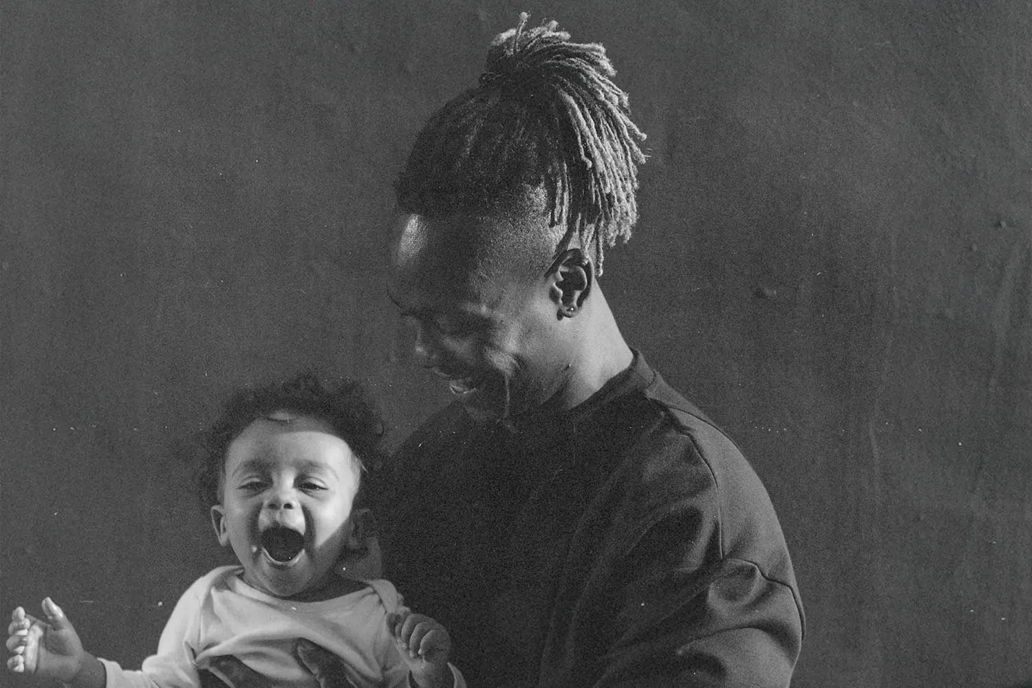A man smiles while holding a small child in his arms in a softly lit, monochrome portrait