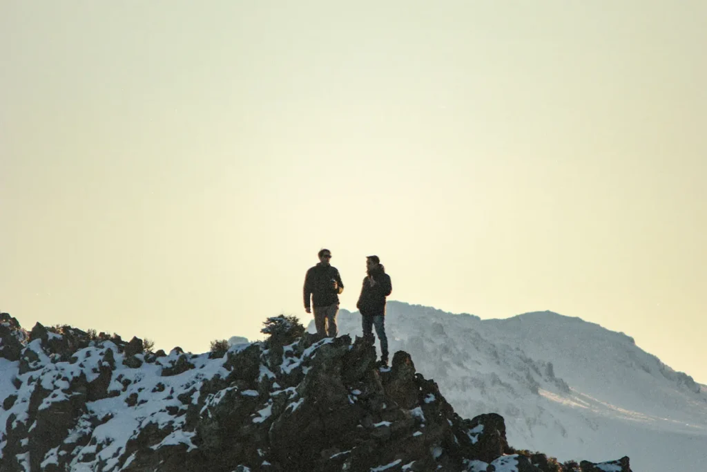 Two people stand on a snowy mountain ridge at sunrise or sunset, silhouetted against a pale sky