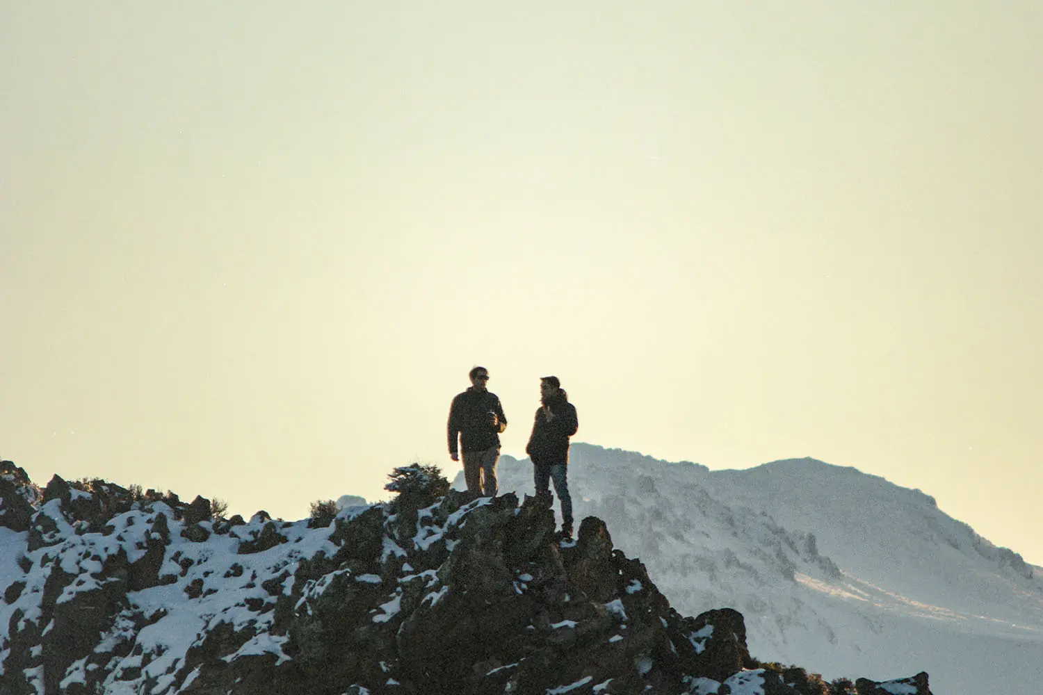 Two people stand on a snowy mountain ridge at sunrise or sunset, silhouetted against a pale sky