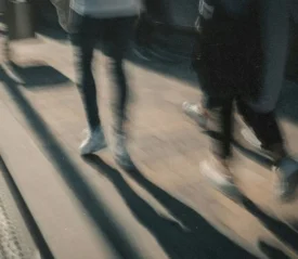 A motion‑blurred view of people walking along a sunlit sidewalk, their long shadows stretching across the ground