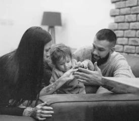 Two adults and a small child sit together on a couch, leaning in as they help the child with something in their hands