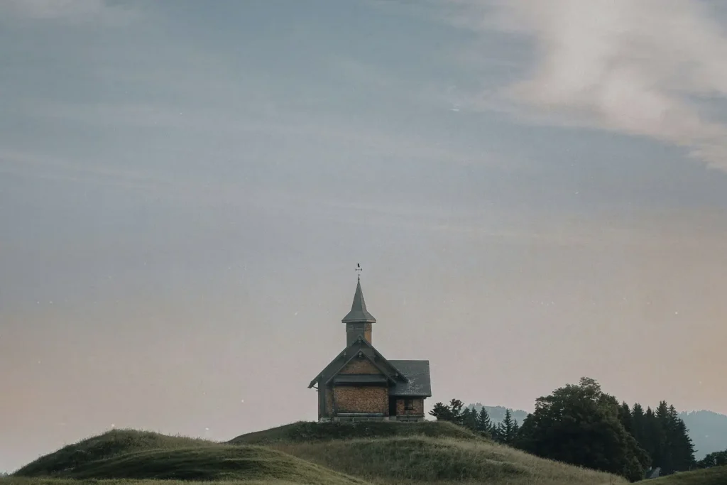 A small wooden chapel with a steeple sits alone on rolling green hills under a soft, pastel sky
