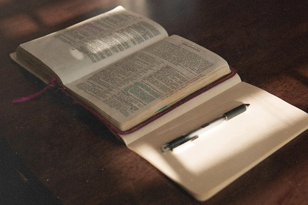 An open Bible lies on a wooden table beside a blank journal and pen, illuminated by soft sunlight