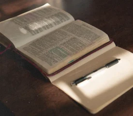 An open Bible lies on a wooden table beside a blank journal and pen, illuminated by soft sunlight