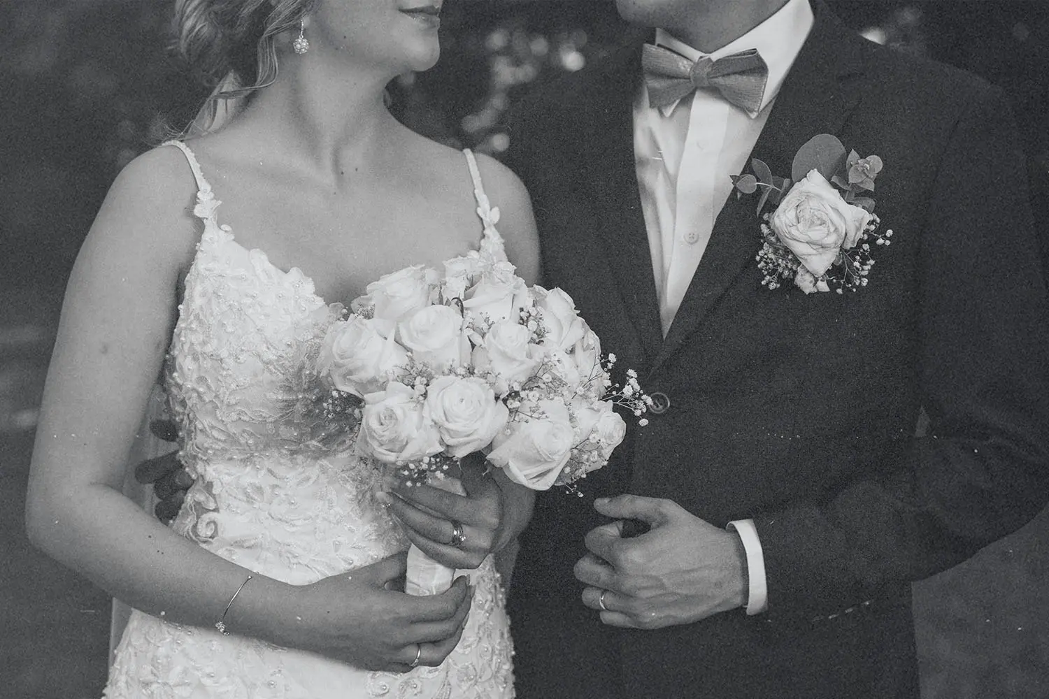 A bride and groom stand close together, the bride holding a bouquet of roses while the groom adjusts his suit jacket