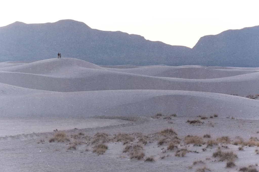 A vast landscape of smooth, rolling sand dunes stretches into the distance, with two people standing together atop a distant dune and a range of dark mountains rising behind them under a pale sky