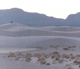 A vast landscape of smooth, rolling sand dunes stretches into the distance, with two people standing together atop a distant dune and a range of dark mountains rising behind them under a pale sky