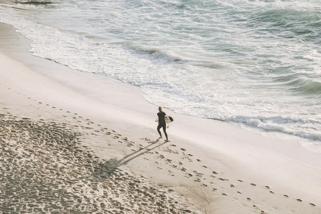 Surfer walking along the shoreline with a board under one arm, leaving footprints in the sand as waves roll in