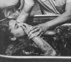 Two people assisting a woman being lowered into a water-filled baptism tub during a baptism ceremony