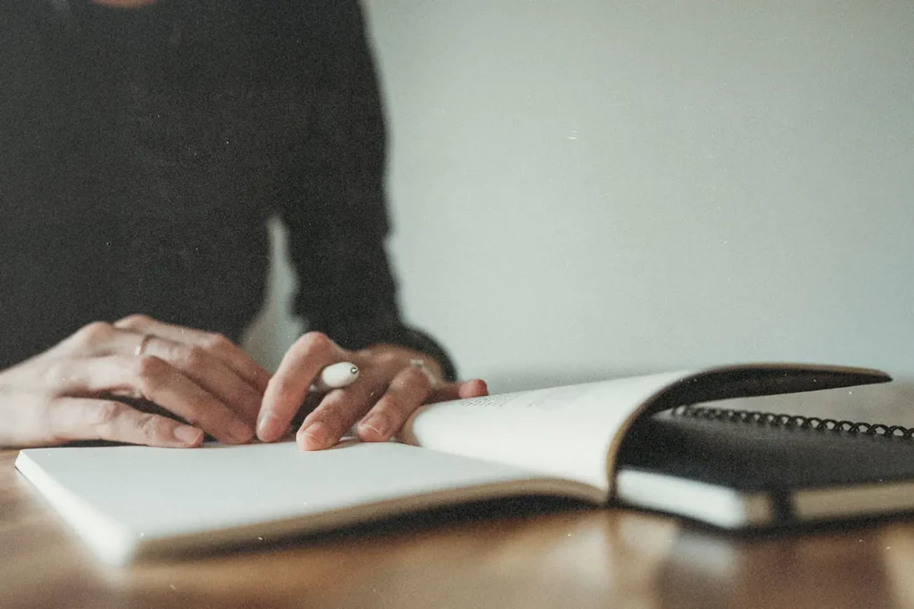 A person writes in an open notebook on a wooden table, with hands resting gently on the page