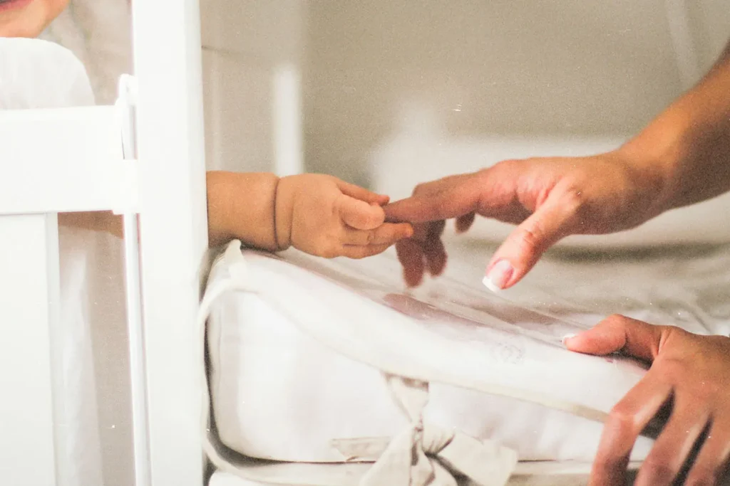 A baby’s hand reaches out from a crib to gently grasp an adult’s fingertip