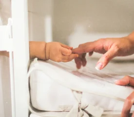 A baby’s hand reaches out from a crib to gently grasp an adult’s fingertip