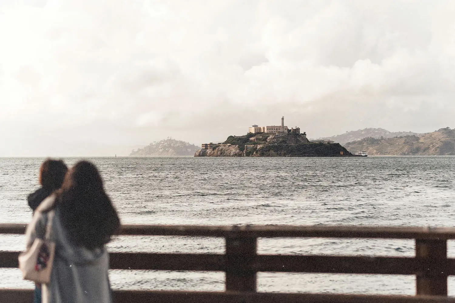 Mother holding her child, standing on a wooden pier looking across the water toward an island with historic stone buildings