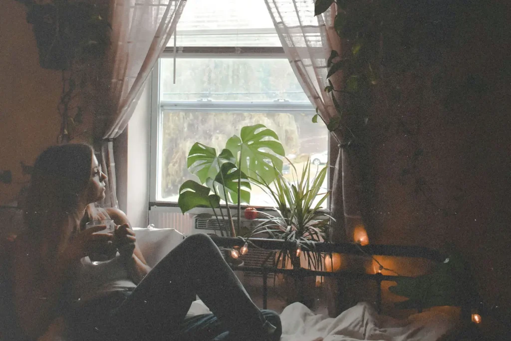 Person sitting on a bed holding a mug while looking out a bright window surrounded by lush indoor plants