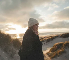 A woman wearing a knit beanie and coat walks along sandy dunes near the beach at sunset, with warm light shining through clouds over the ocean