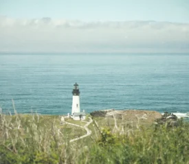 A white lighthouse stands on a grassy cliff overlooking calm blue ocean water, with clouds lining the distant horizon
