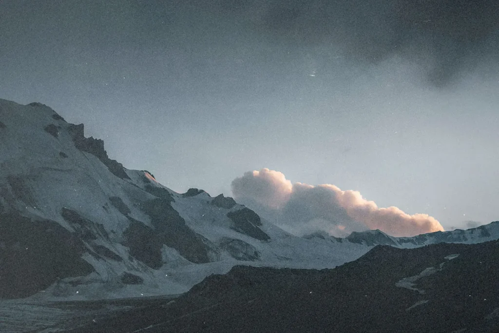Snow‑covered mountain peaks rise sharply into the sky, illuminated by soft light with a cluster of clouds hovering above
