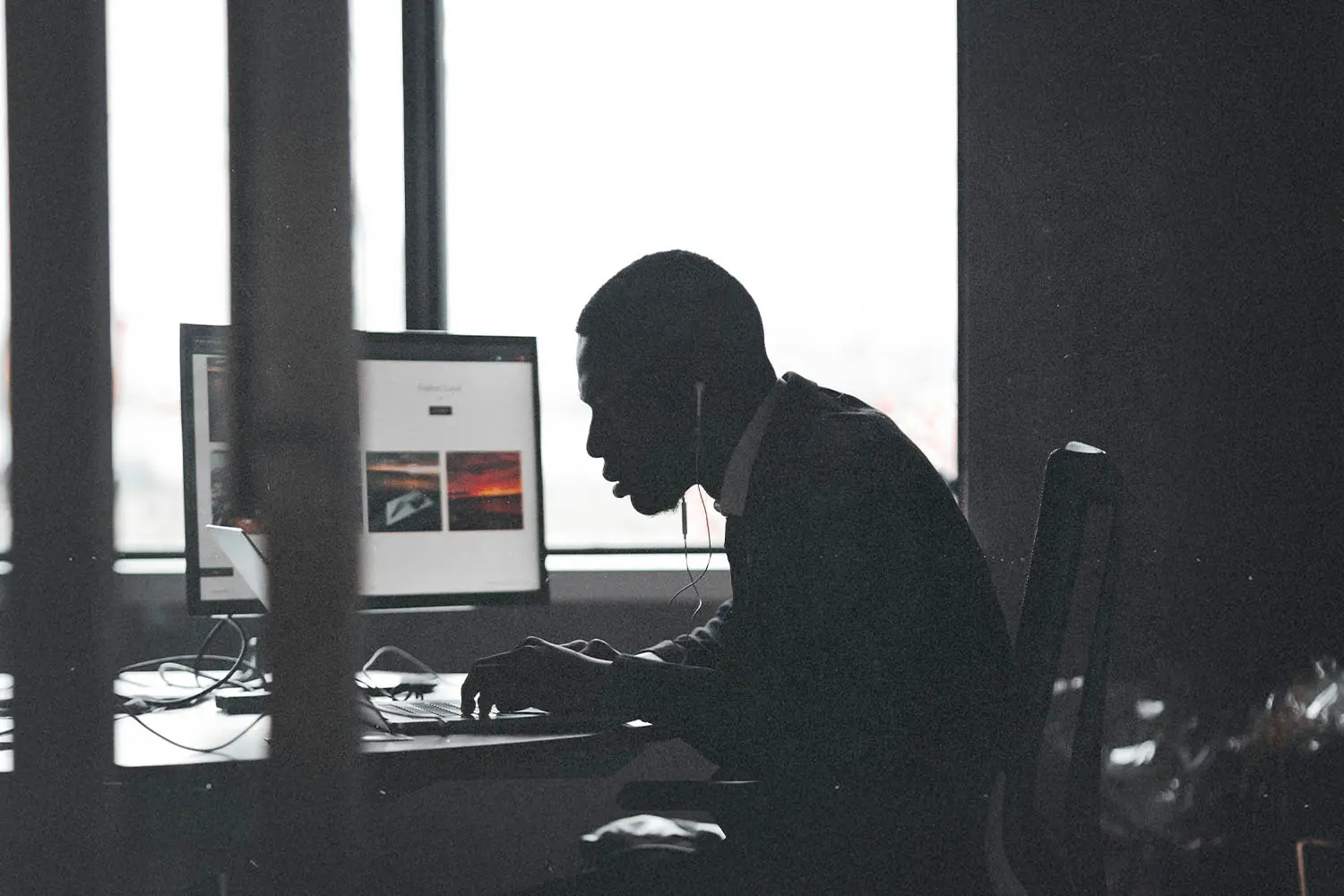 A person sits at a desk working on a computer with large windows behind them, silhouetted against the light