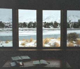Open Bibles on a wooden table inside a cabin, overlooking a snowy landscape and frozen river through large windows