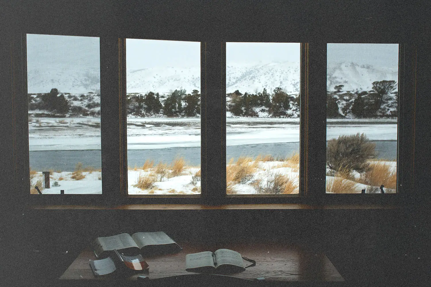 Open Bibles on a wooden table inside a cabin, overlooking a snowy landscape and frozen river through large windows
