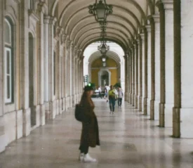 Person stands in a long arched hallway with stone columns and hanging lanterns, while others walk in the distance