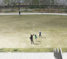 Two children run and play on a large open grassy field while an adult watches nearby