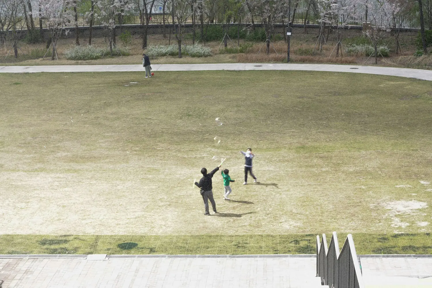 Two children run and play on a large open grassy field while an adult watches nearby