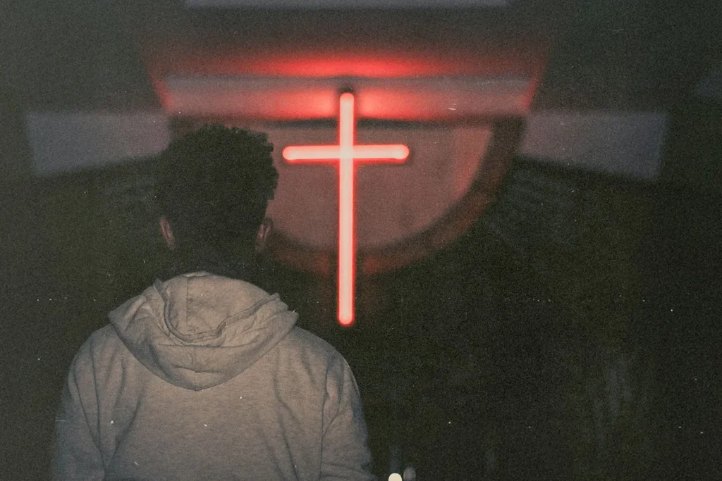Silhouetted person standing in a dark room facing a glowing red cross on the wall
