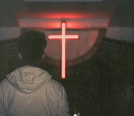 Silhouetted person standing in a dark room facing a glowing red cross on the wall