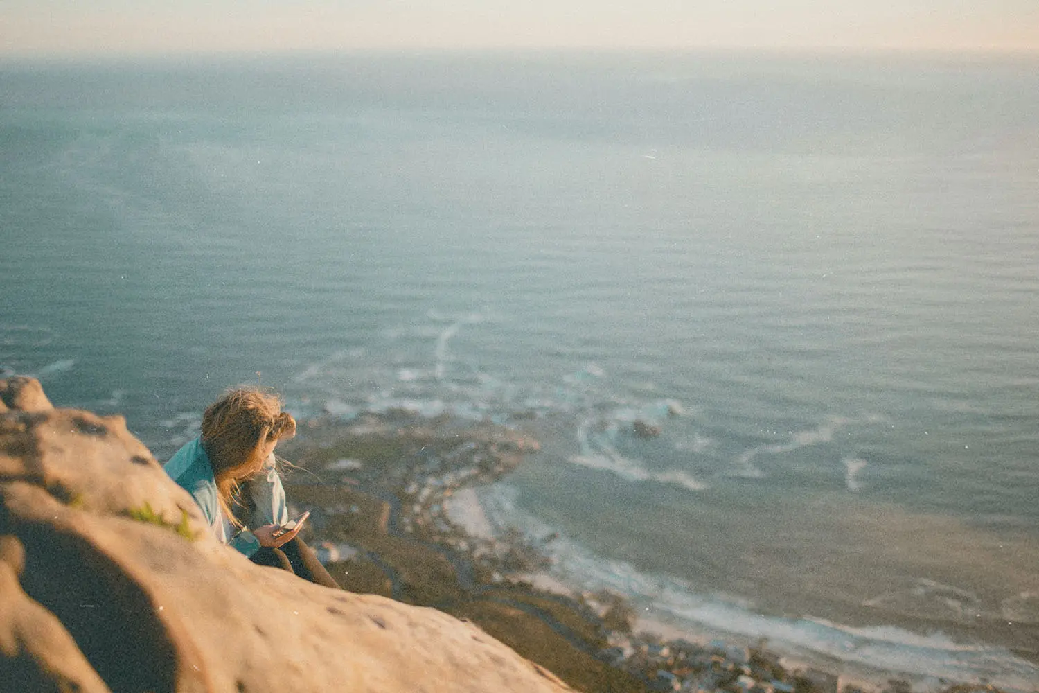 Person sitting on a rocky cliff overlooking the ocean at sunset, with waves and coastline visible far below
