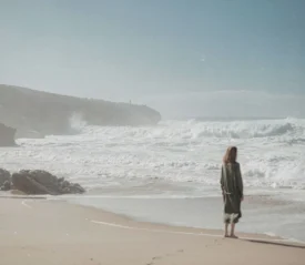 Person standing alone on a sandy beach, watching waves crash against a rocky shoreline under a clear blue sky