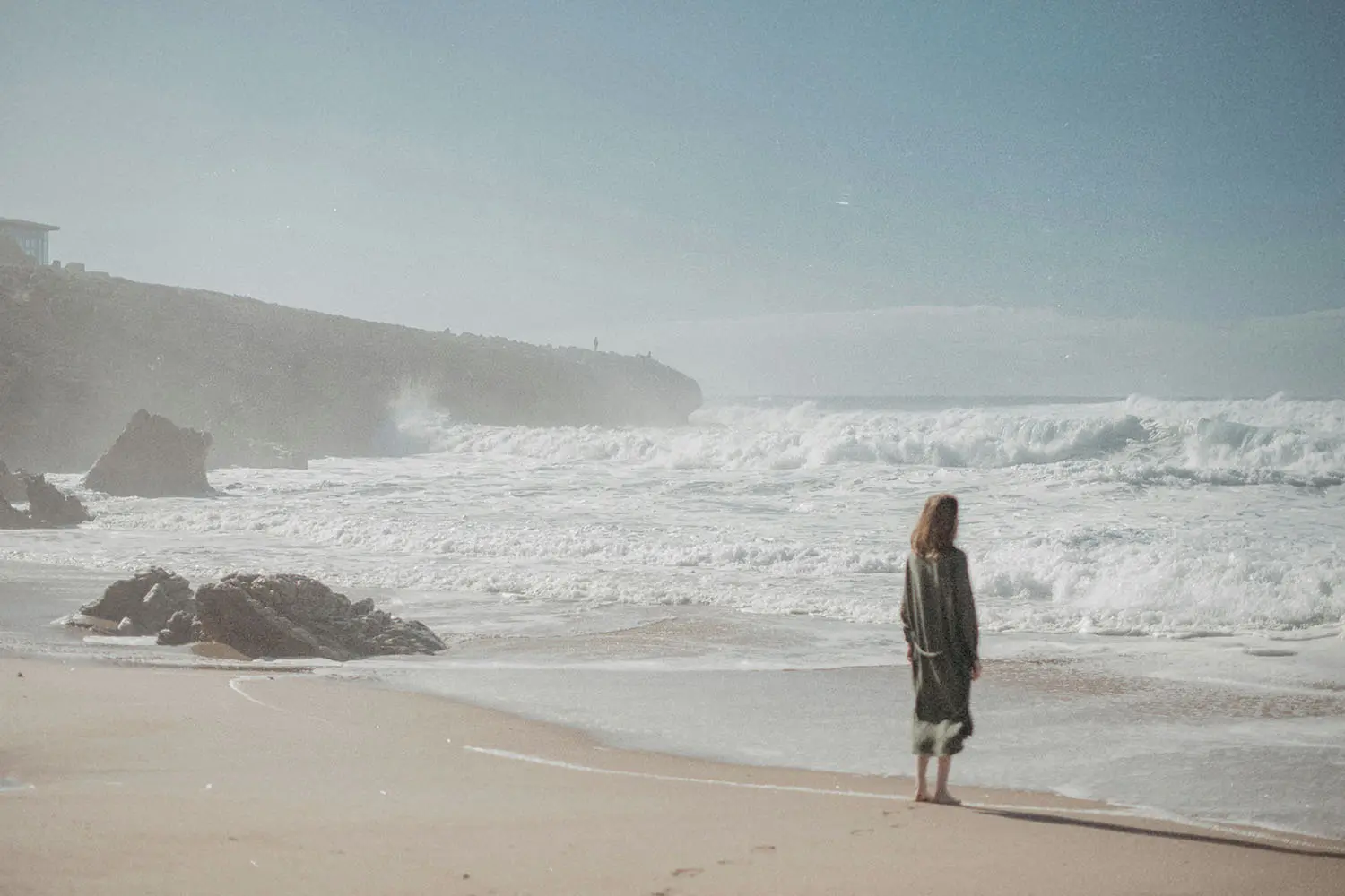 Person standing alone on a sandy beach, watching waves crash against a rocky shoreline under a clear blue sky