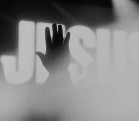 Silhouetted hand raised toward an illuminated screen with "Jesus" in bold text in the background
