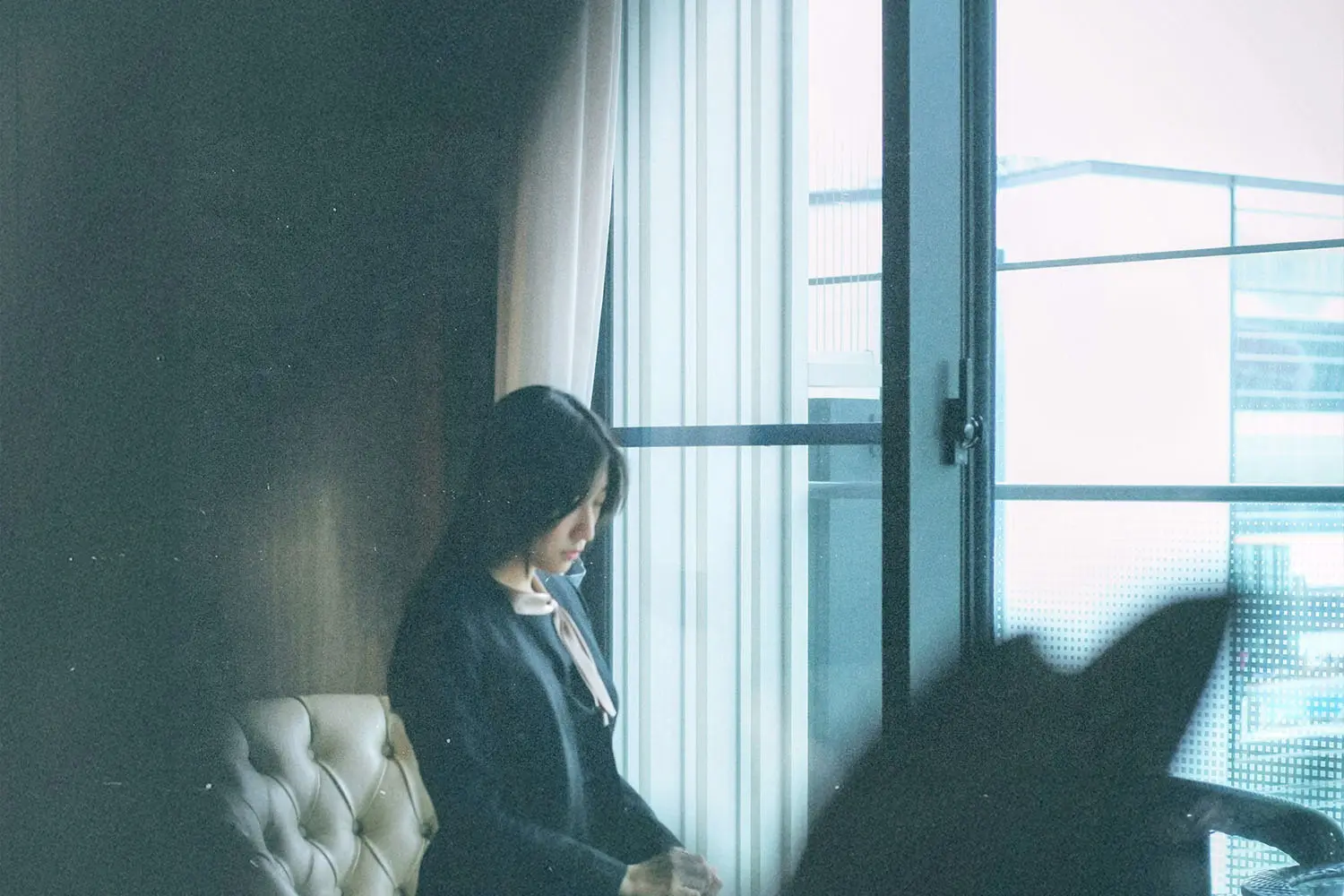 Person sitting in a softly lit room, looking out a large window with sheer curtains and an urban view outside
