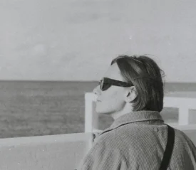 Person standing at a waterfront railing, looking out toward the open sea on a calm, overcast day